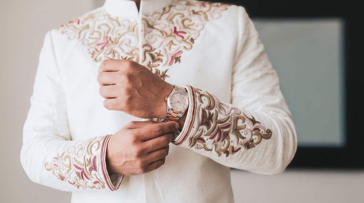 Man in ornate white and gold embroidered sherwani adjusting cufflinks, traditional Indian wedding attire.