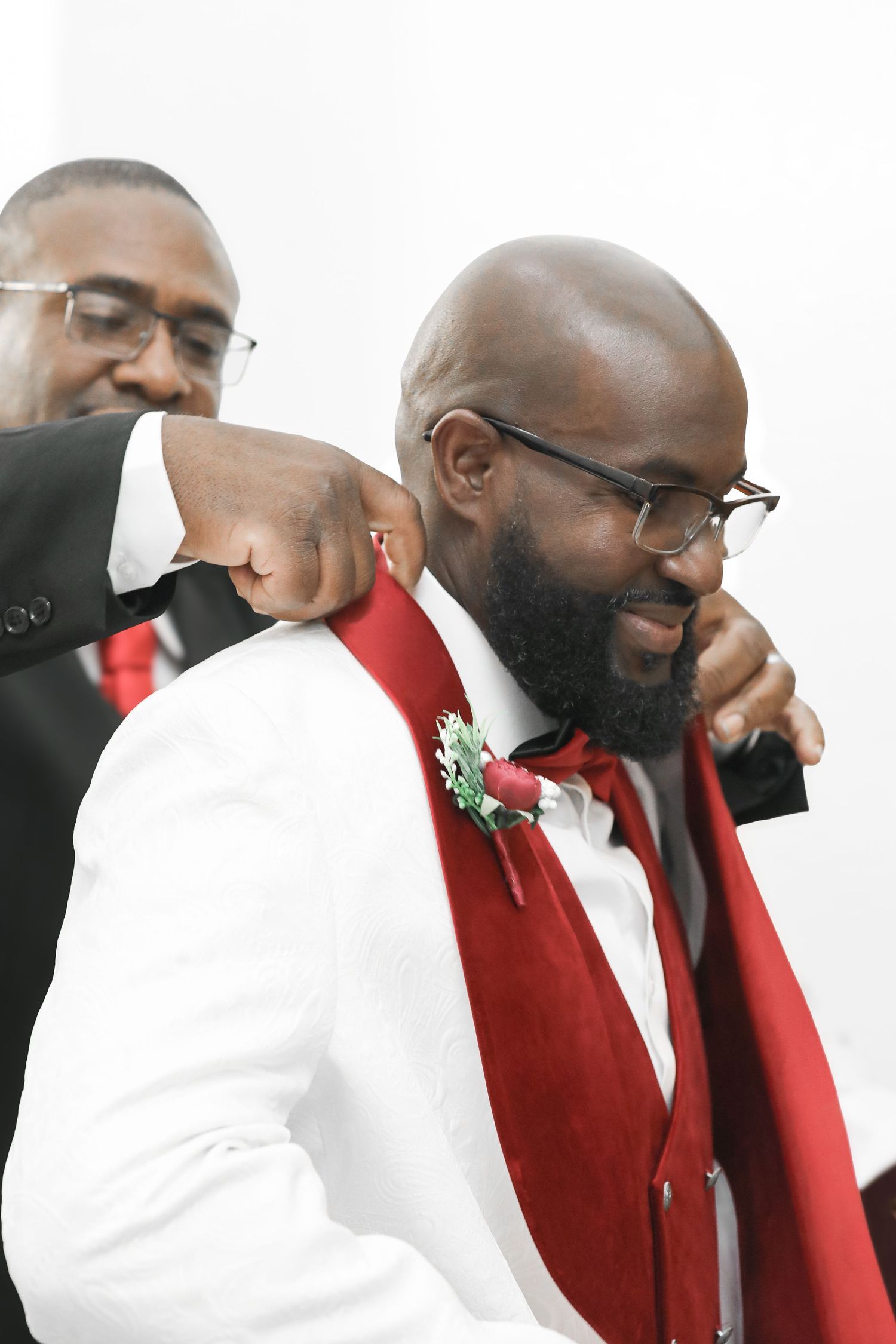 An emotional moment at a wedding shows a man in a white suit with red vest being comforted by another in a tender embrace.