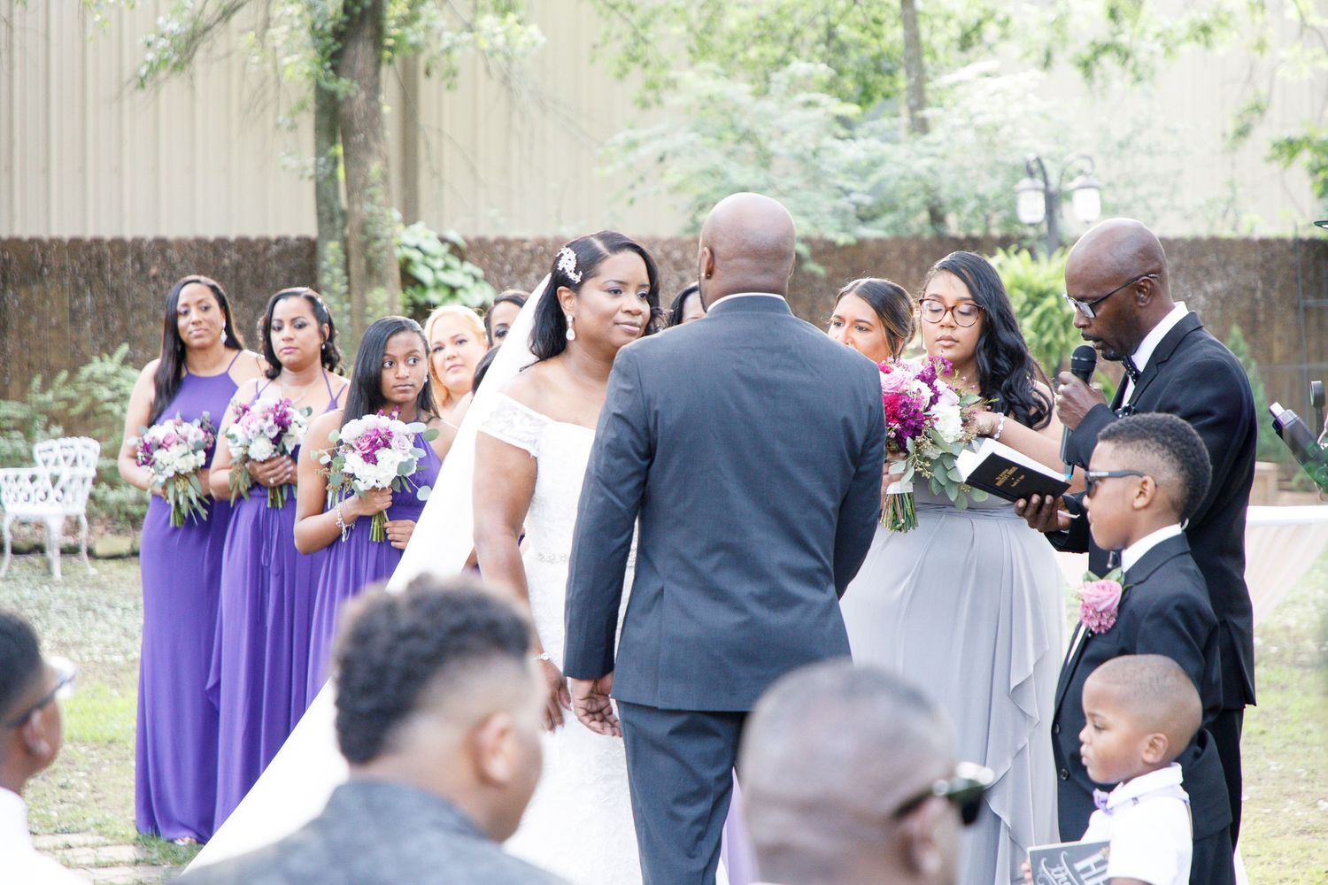 Outdoor wedding ceremony with bride and groom at altar, bridesmaids in purple dresses, and officiant reading vows.