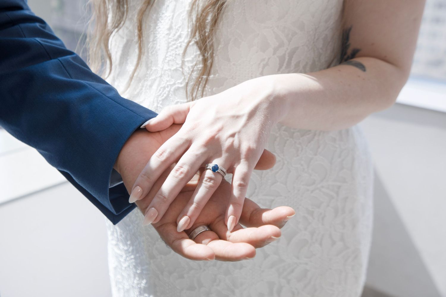 Bride and groom holding hands showing wedding rings, bride wearing white lace dress with sapphire engagement ring.