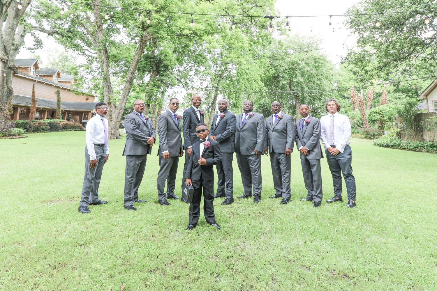 Groomsmen in gray suits pose outdoors on green lawn with trees and building backdrop at wedding celebration.