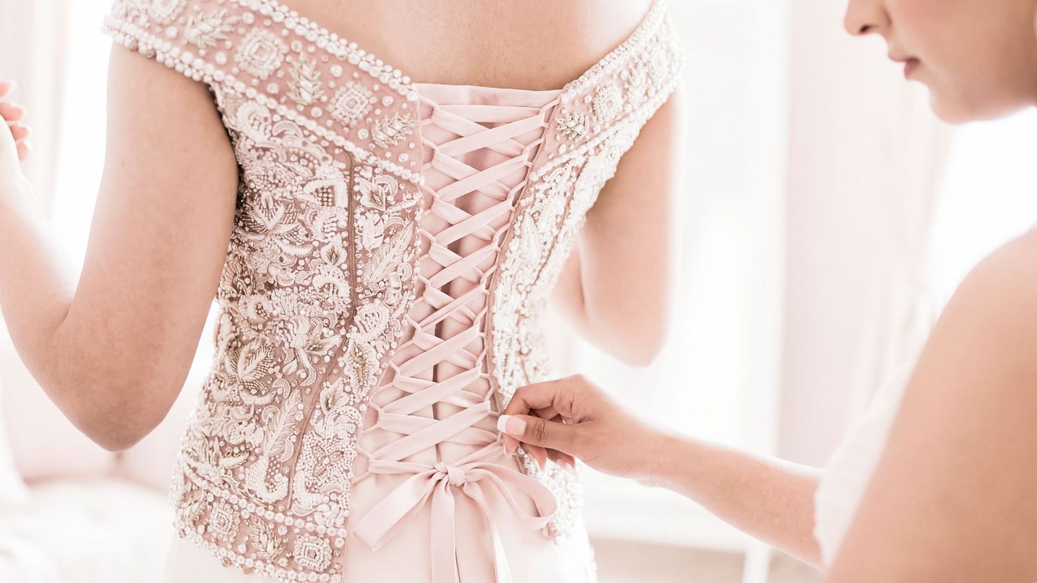 A bride in a blush pink beaded corset wedding gown being laced up by a bridesmaid in a bright white room.