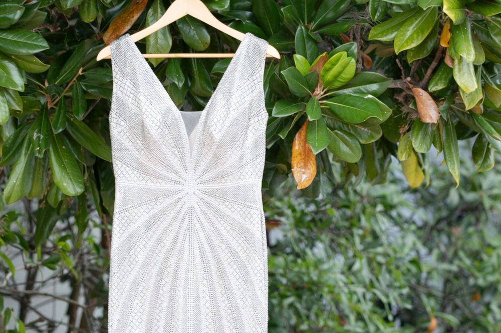 White lace wedding dress on wooden hanger displayed against lush green magnolia tree foliage outdoors.