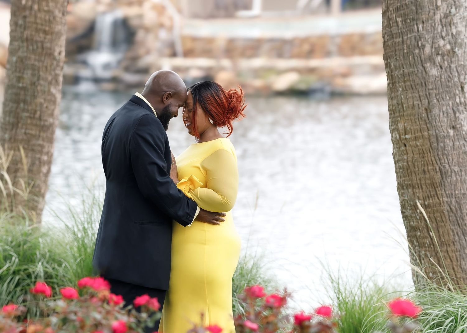 A couple embraces romantically in a garden setting, woman in yellow dress, near a fountain with pink flowers.
