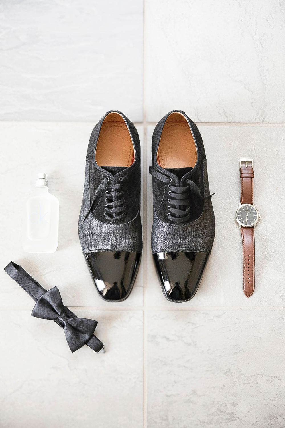 Black cap-toe oxford dress shoes displayed with a brown leather watch, black bow tie, and cologne bottle on marble floor.