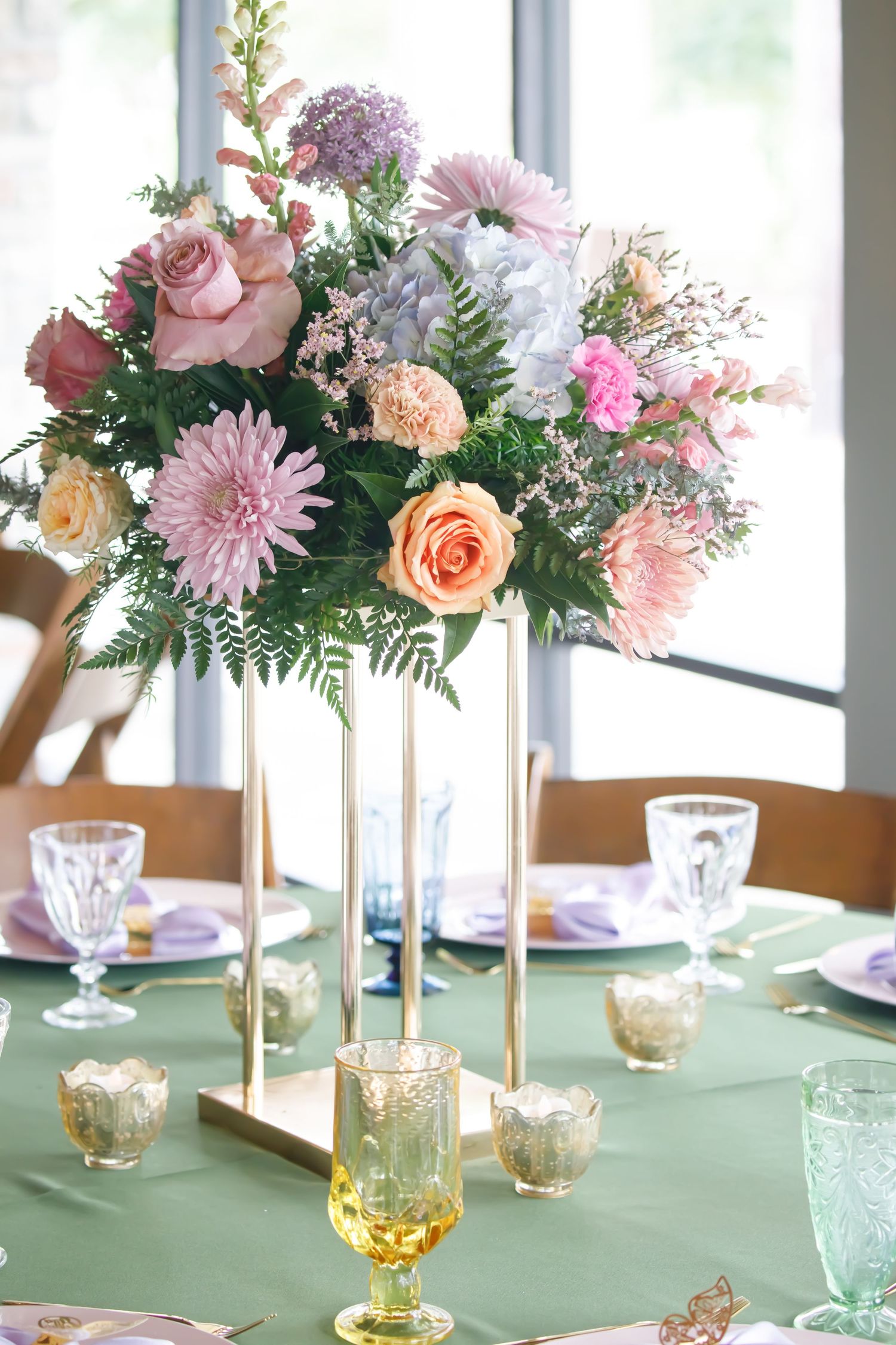 Elegant floral centerpiece with pink roses and dahlias on gold stands atop a sage green tablecloth with vintage glassware.