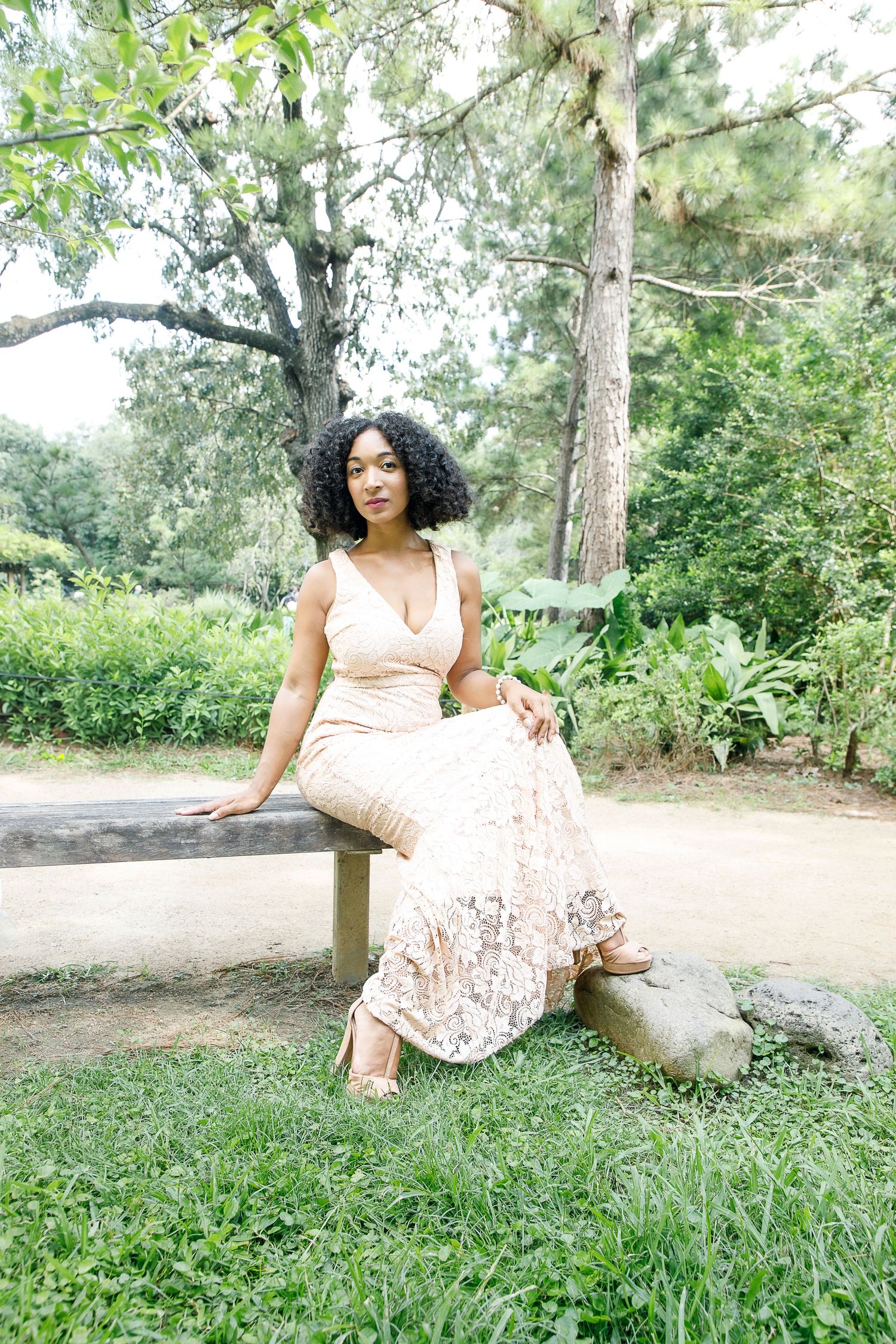 A person in a pale pink lace dress poses gracefully on a fallen tree trunk in a serene outdoor garden setting.