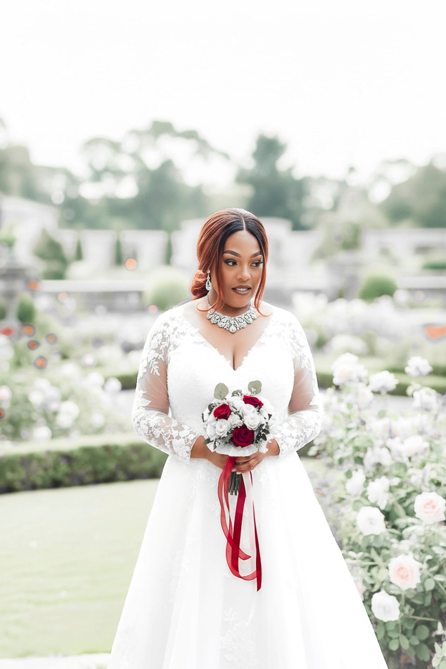 Bride in elegant white lace wedding dress holds burgundy and white floral bouquet in outdoor garden setting.