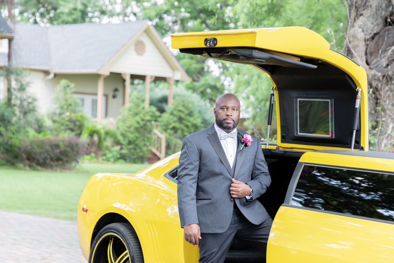Man in gray suit poses beside a yellow sports car with open trunk door outdoors.
