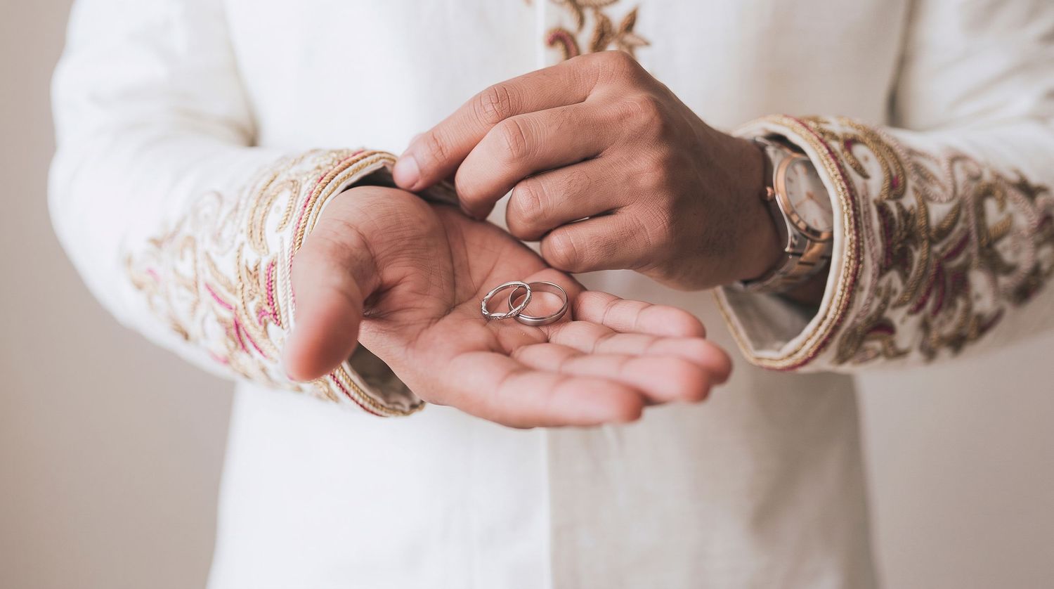 A groom in traditional white embroidered attire holds wedding rings in his palm during an Indian wedding ceremony.