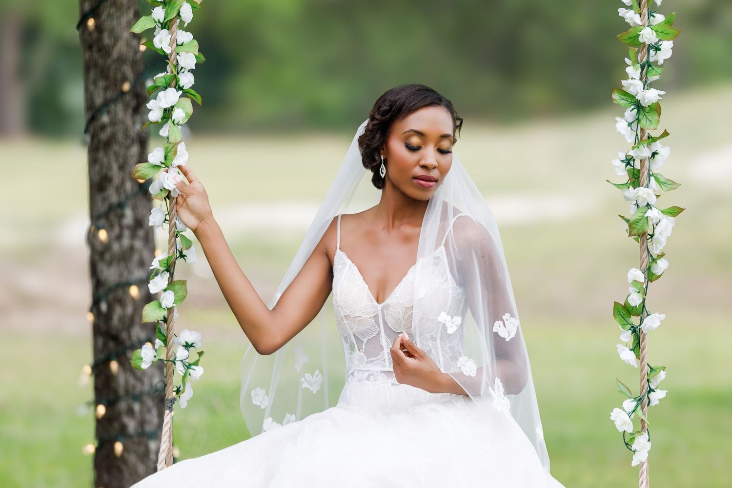 Elegant Black bride in a white lace wedding gown and veil sits on a floral swing outdoors.
