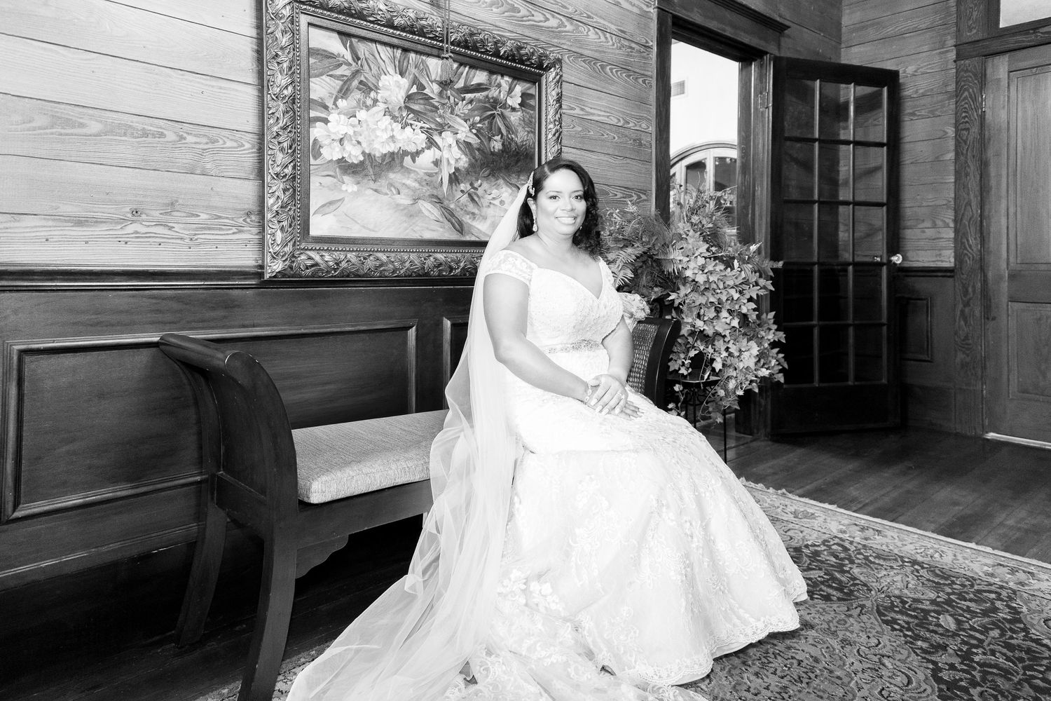Black and white bridal portrait of a woman in a white wedding gown seated on a bench in an elegant wood-paneled room.