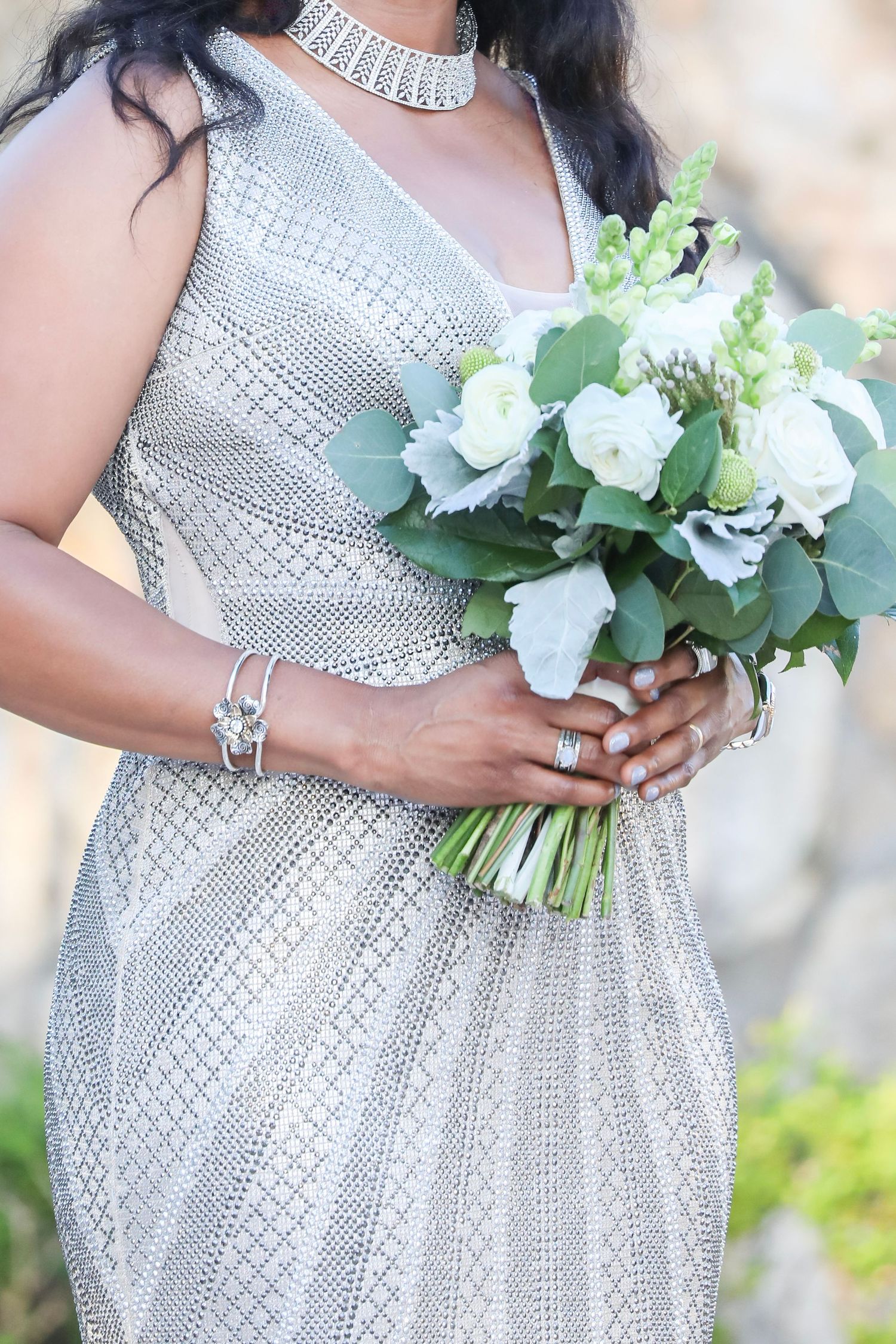 Close-up of silver sequined dress with crystal necklace and white floral bouquet held by hands adorned with bracelet.