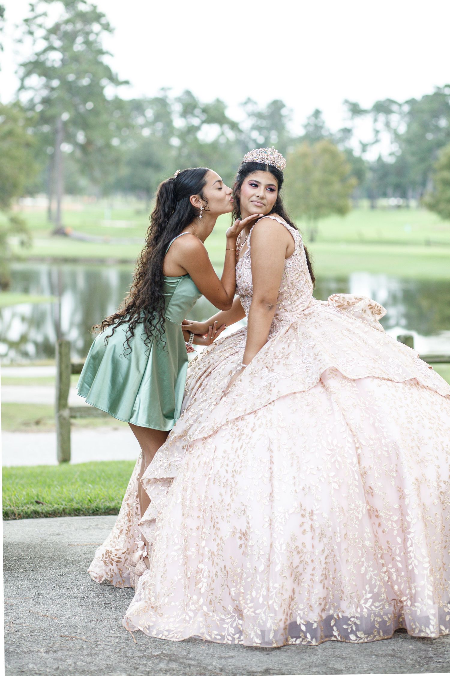 Two people pose together outdoors in formal attire with one wearing a flowing pink ballgown on grassy grounds.