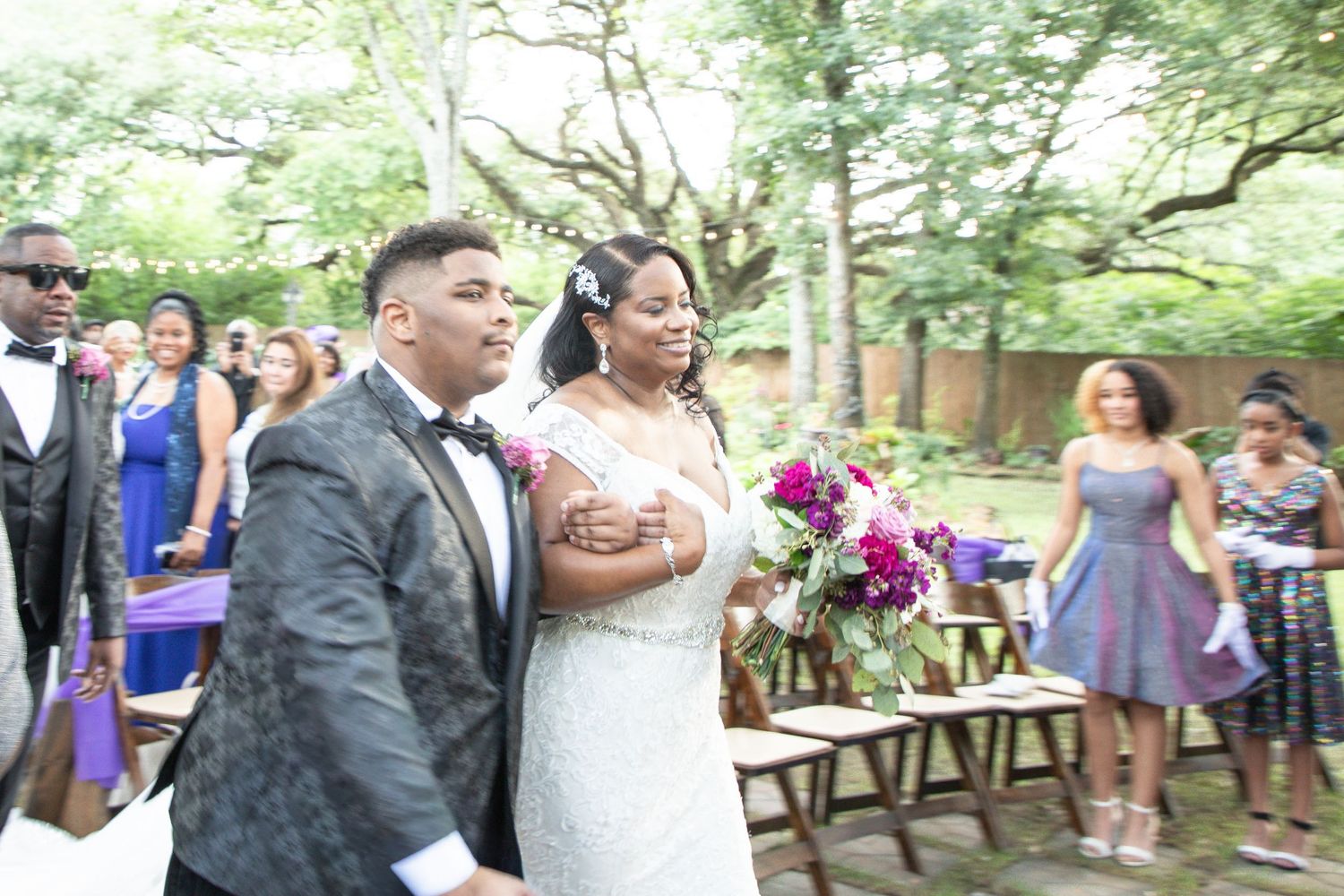 Bride in white lace gown holding purple bouquet walks down outdoor aisle with escort at elegant garden wedding ceremony.