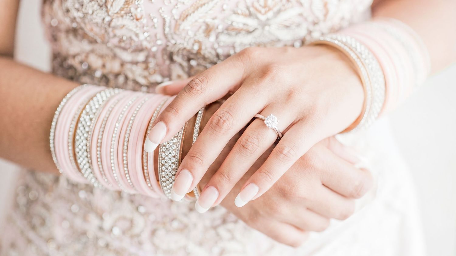 Bride wearing pink bangles and a diamond engagement ring on her manicured hands in a white lace dress.