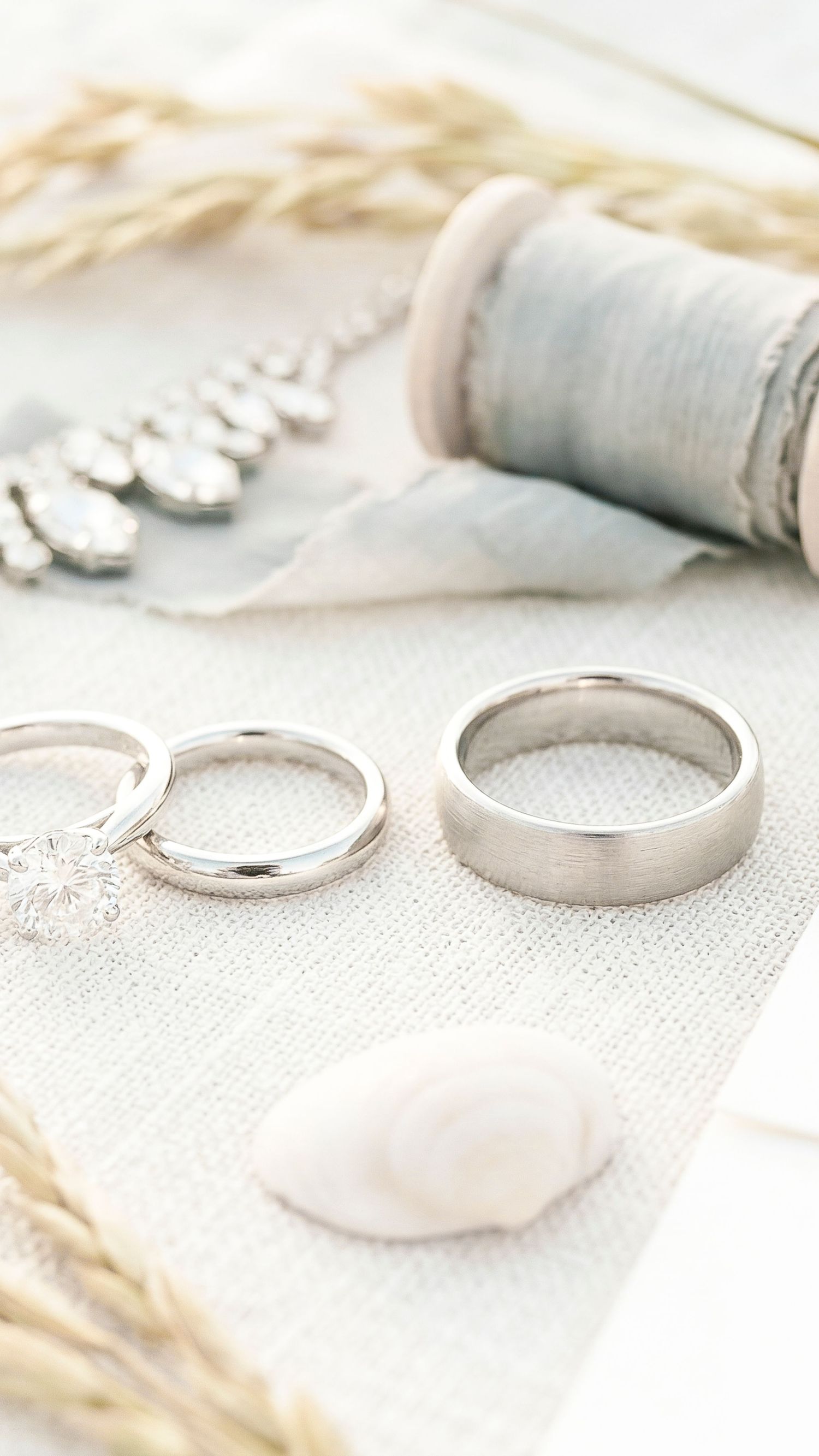 Two silver wedding bands displayed on linen with a spool of thread, dried flowers, and crystal jewelry.
