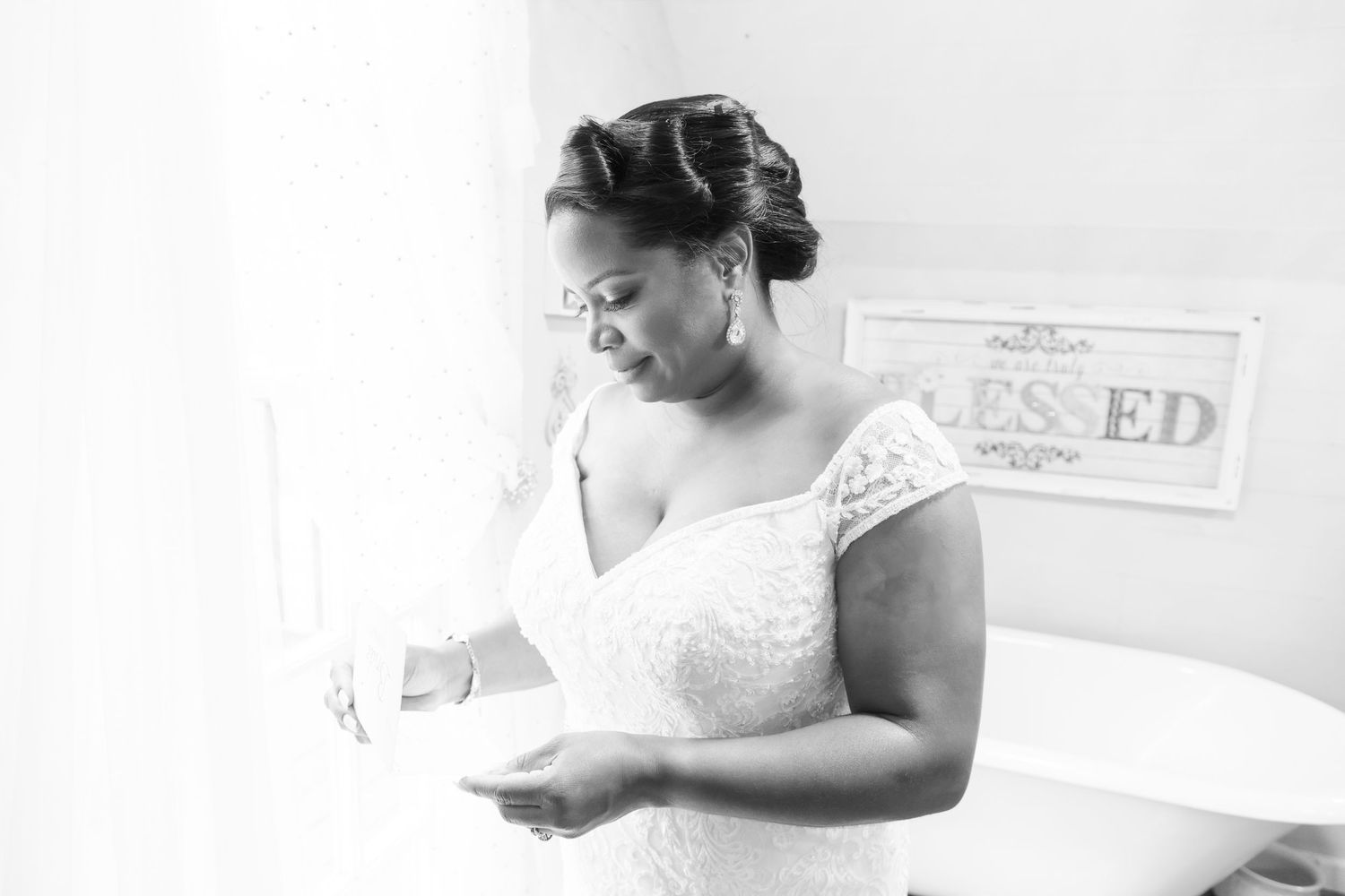 Black and white photo of a bride in lace gown reading a letter near a window on her wedding day.