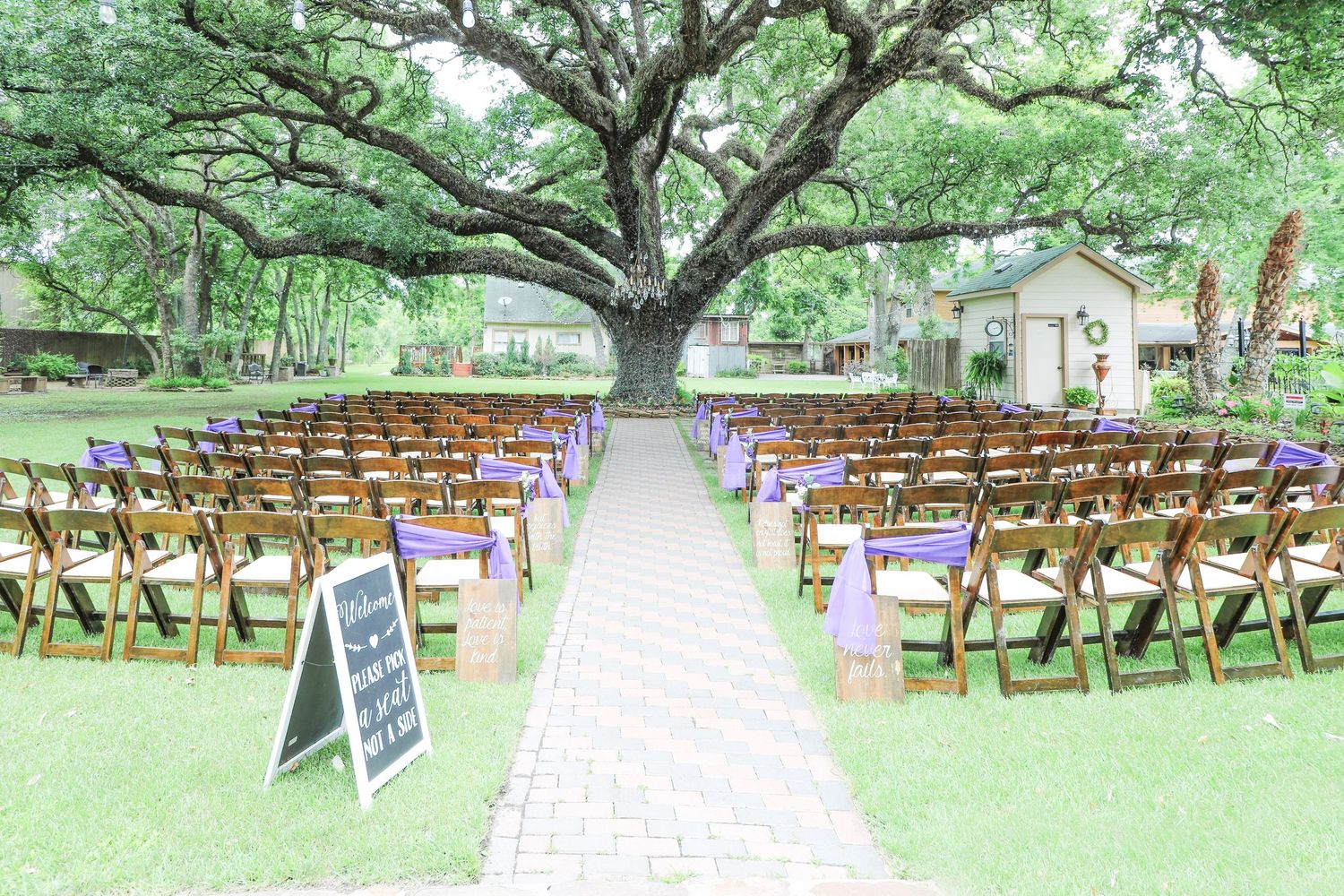 Outdoor wedding ceremony setup with wooden chairs, purple ribbon accents, brick aisle, and large oak tree backdrop.