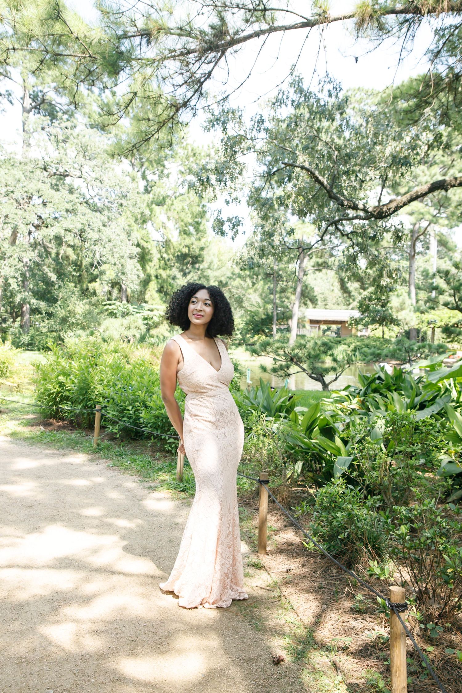 A person in a white summer dress walks along a sunlit garden path surrounded by lush greenery and trees.