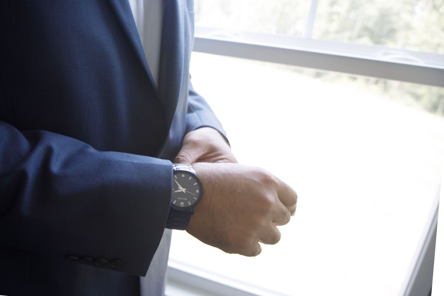 Man in navy blue suit adjusting wristwatch near window, wearing elegant black analog watch.