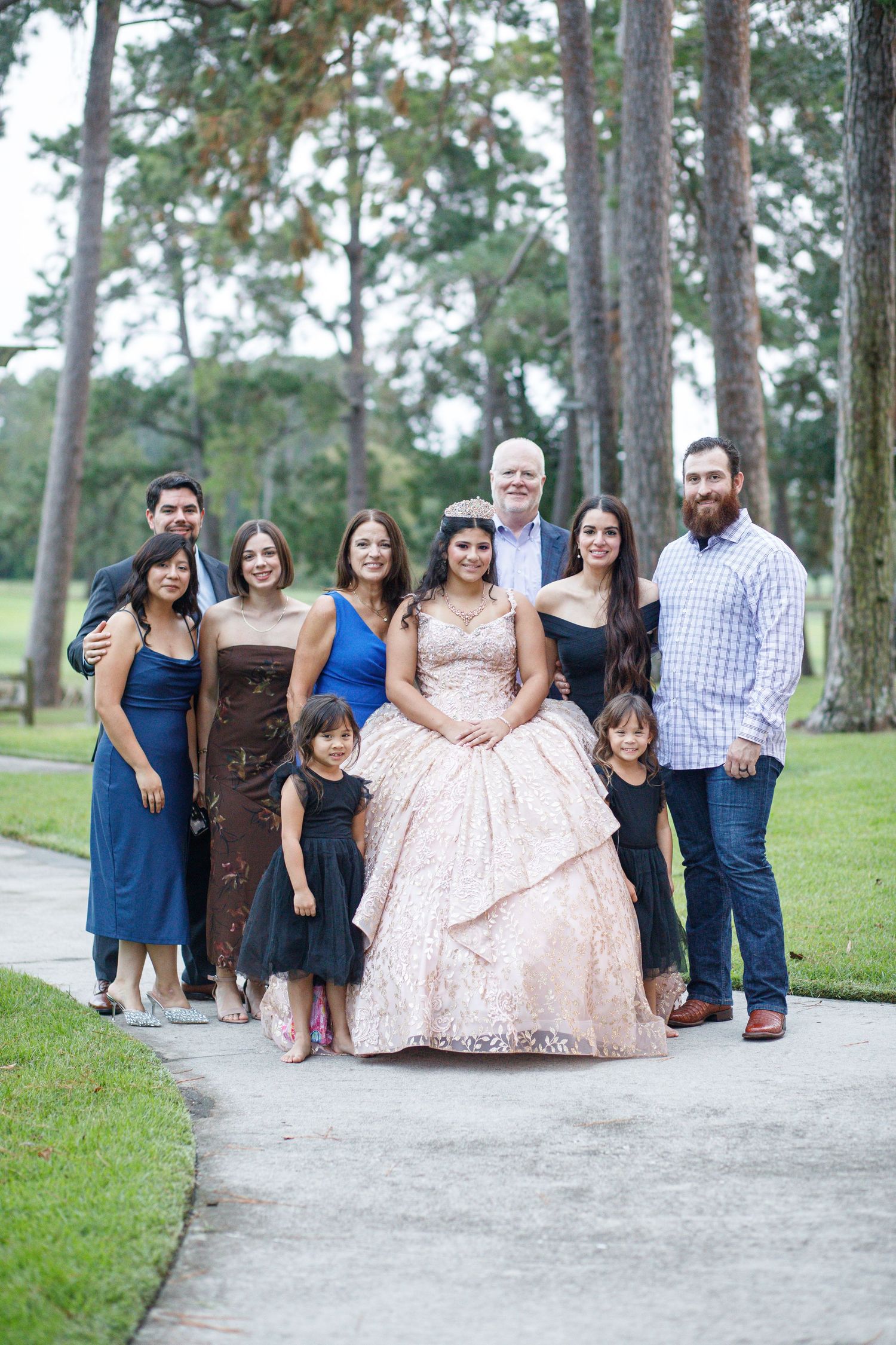 A group of people wearing formal attire pose together on a pathway surrounded by tall pine trees in a park setting.