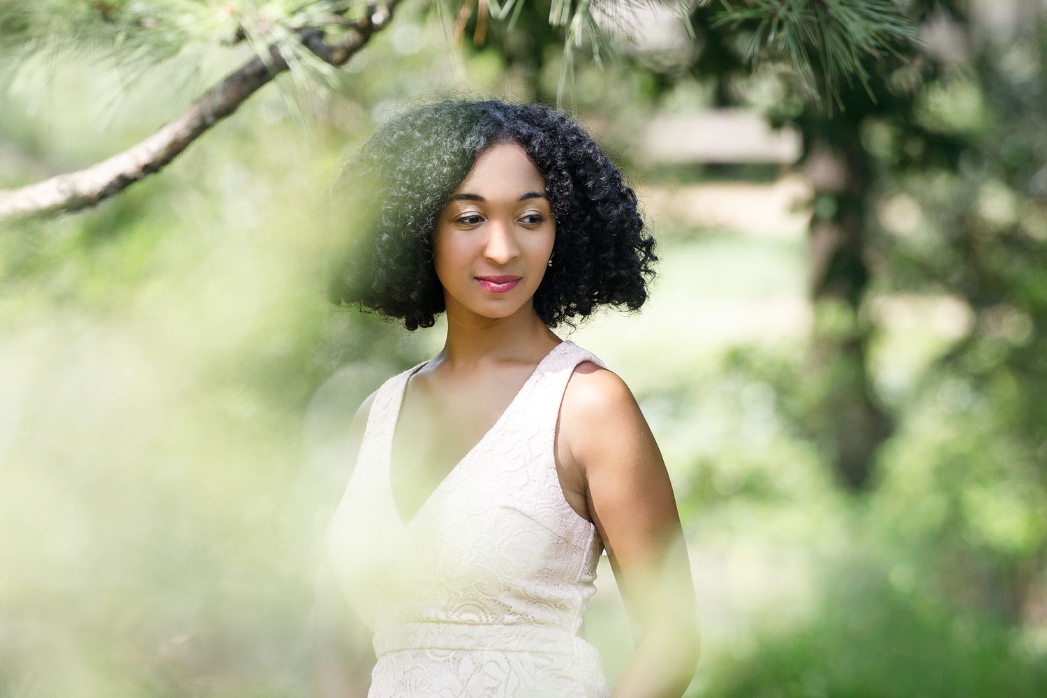 A person with curly black hair poses outdoors in natural lighting among green foliage.