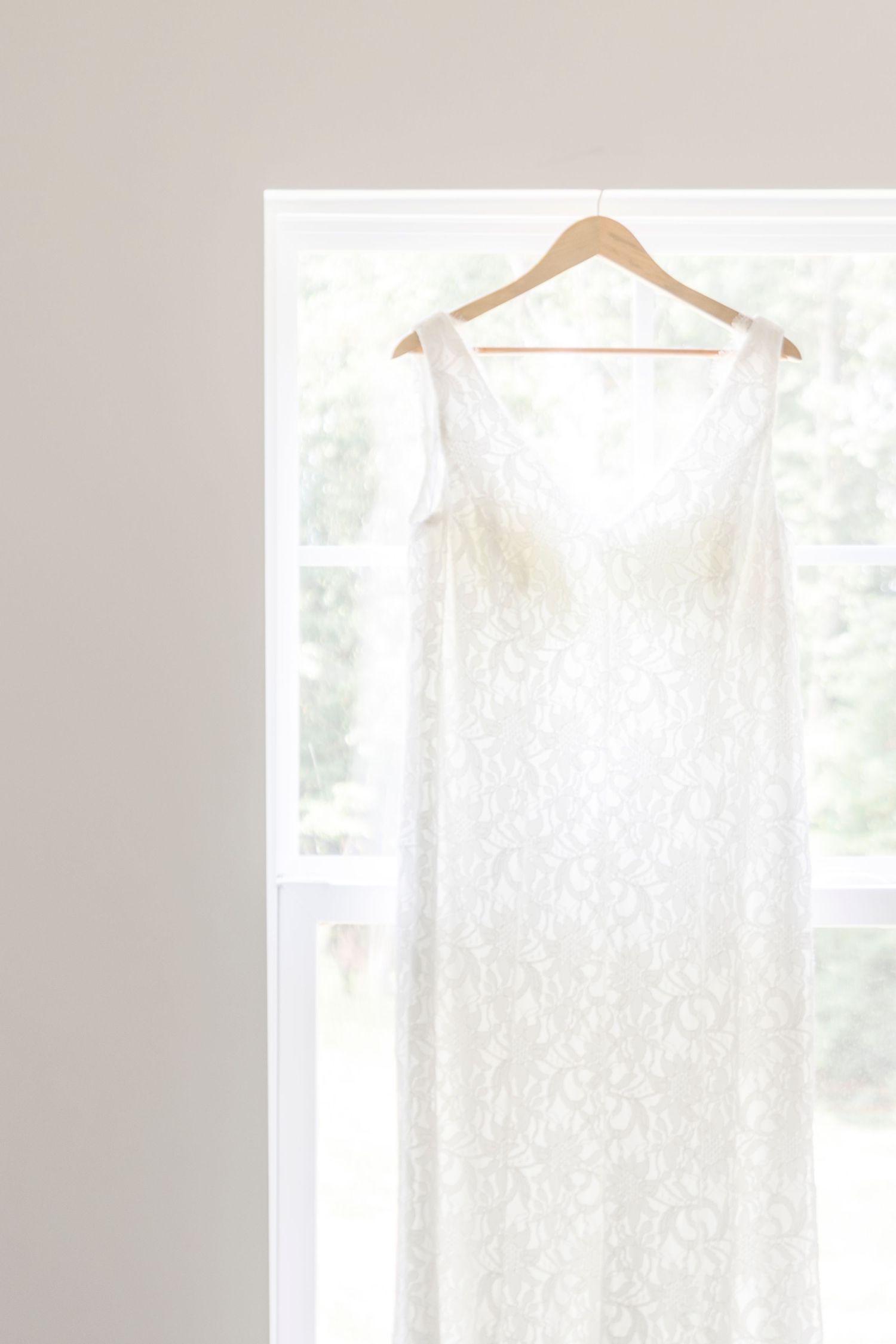 A delicate white lace wedding dress hanging on a wooden hanger in a bright window doorway.