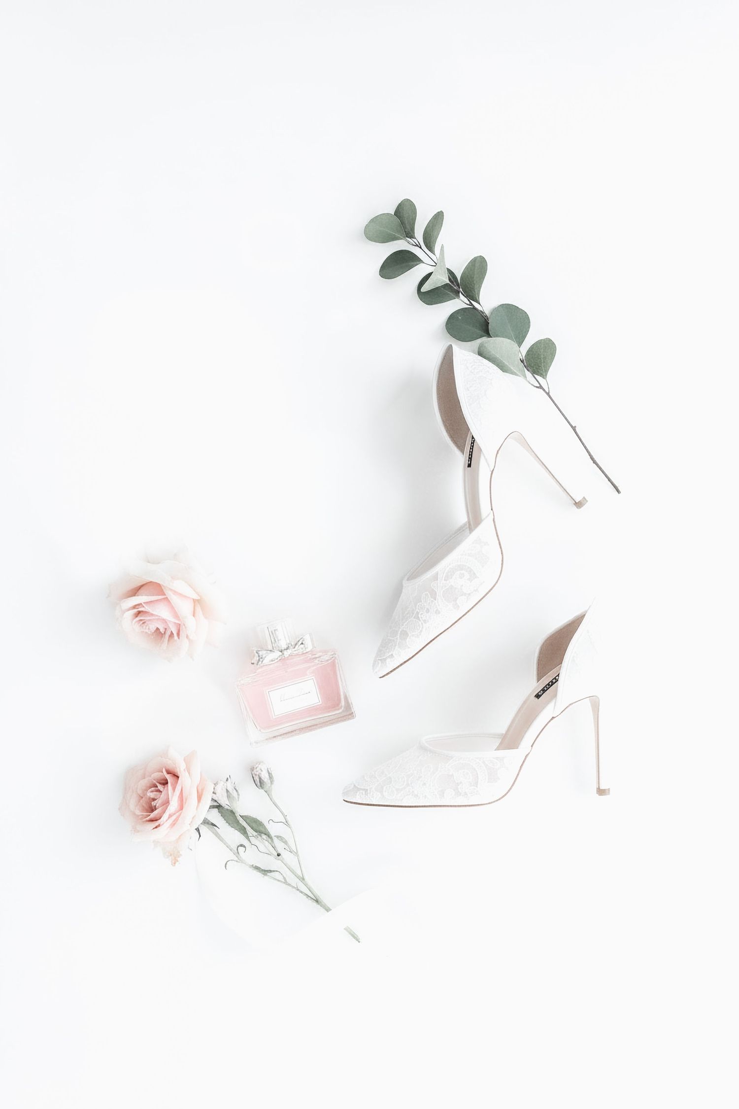White lace bridal heels with pink roses, eucalyptus sprigs, and pink perfume bottle on white background.