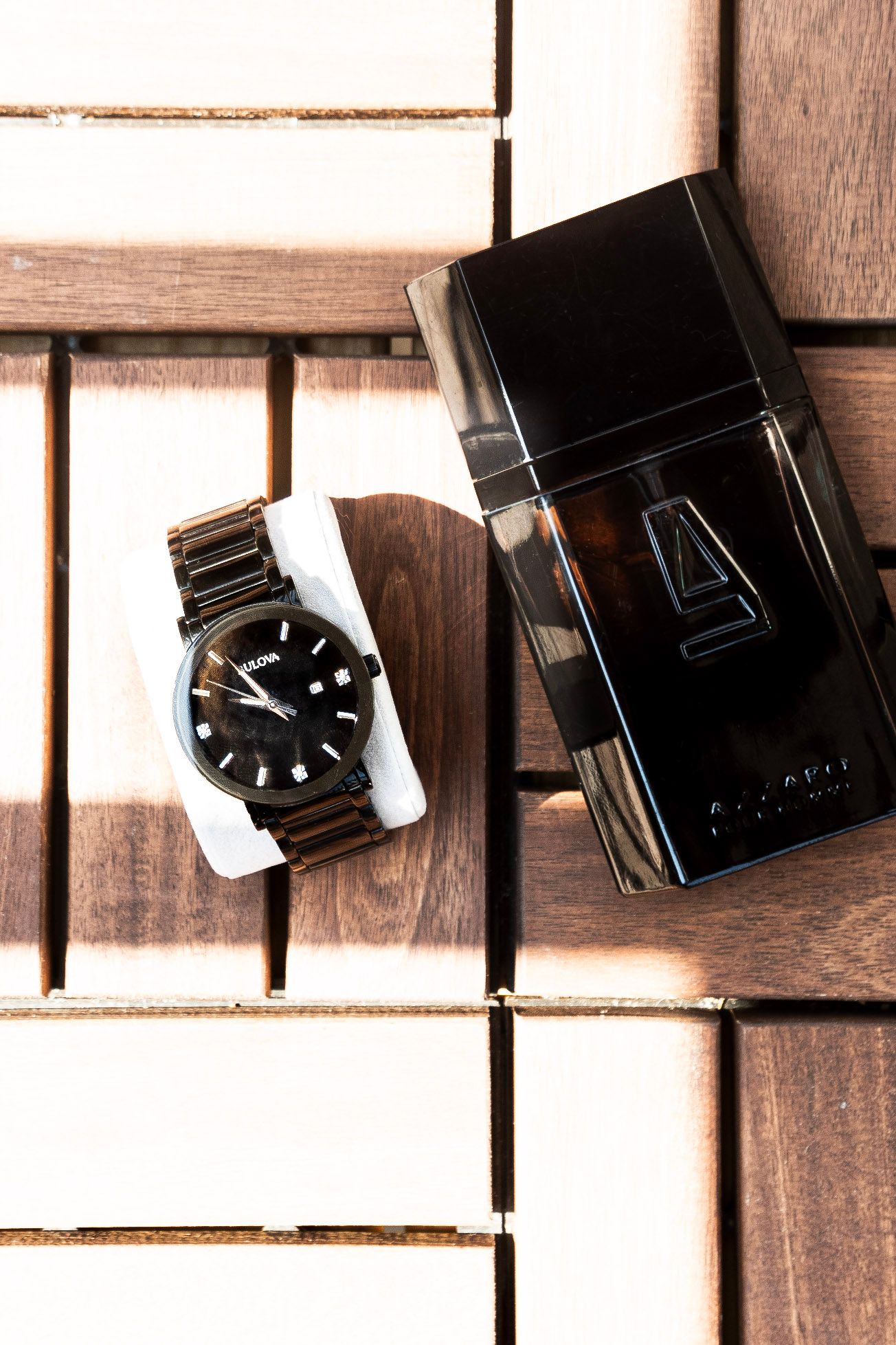 Black men's watch and dark cologne bottle displayed on wooden slat surface in natural light.