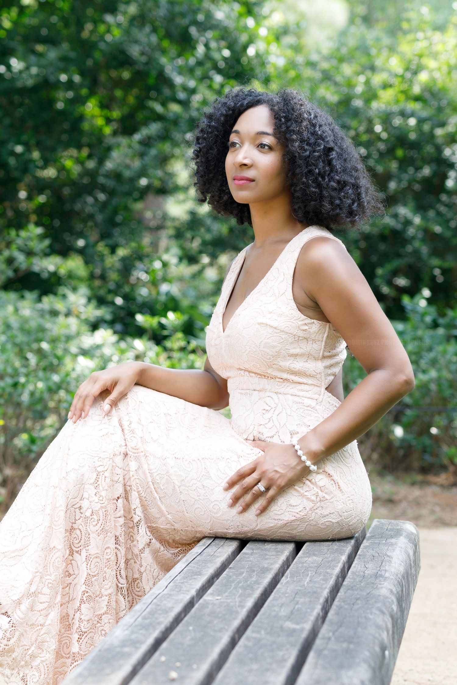A person in a peach lace dress poses gracefully on a wooden bench in an outdoor garden setting.