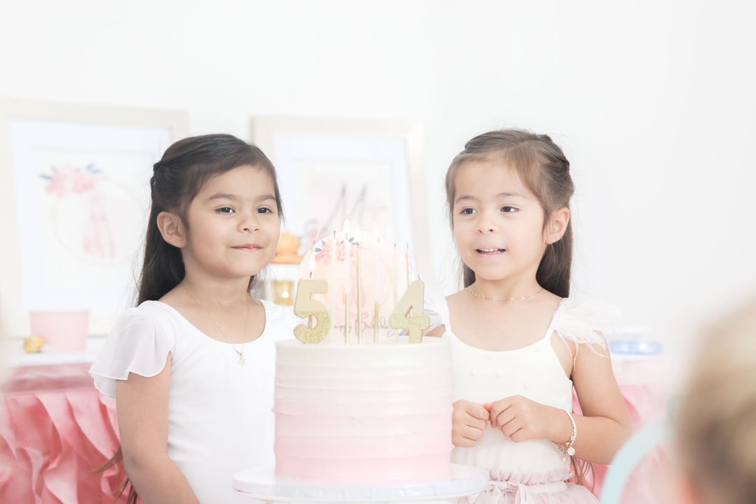 Children in pink dresses gather around a birthday cake at a festive party celebration.