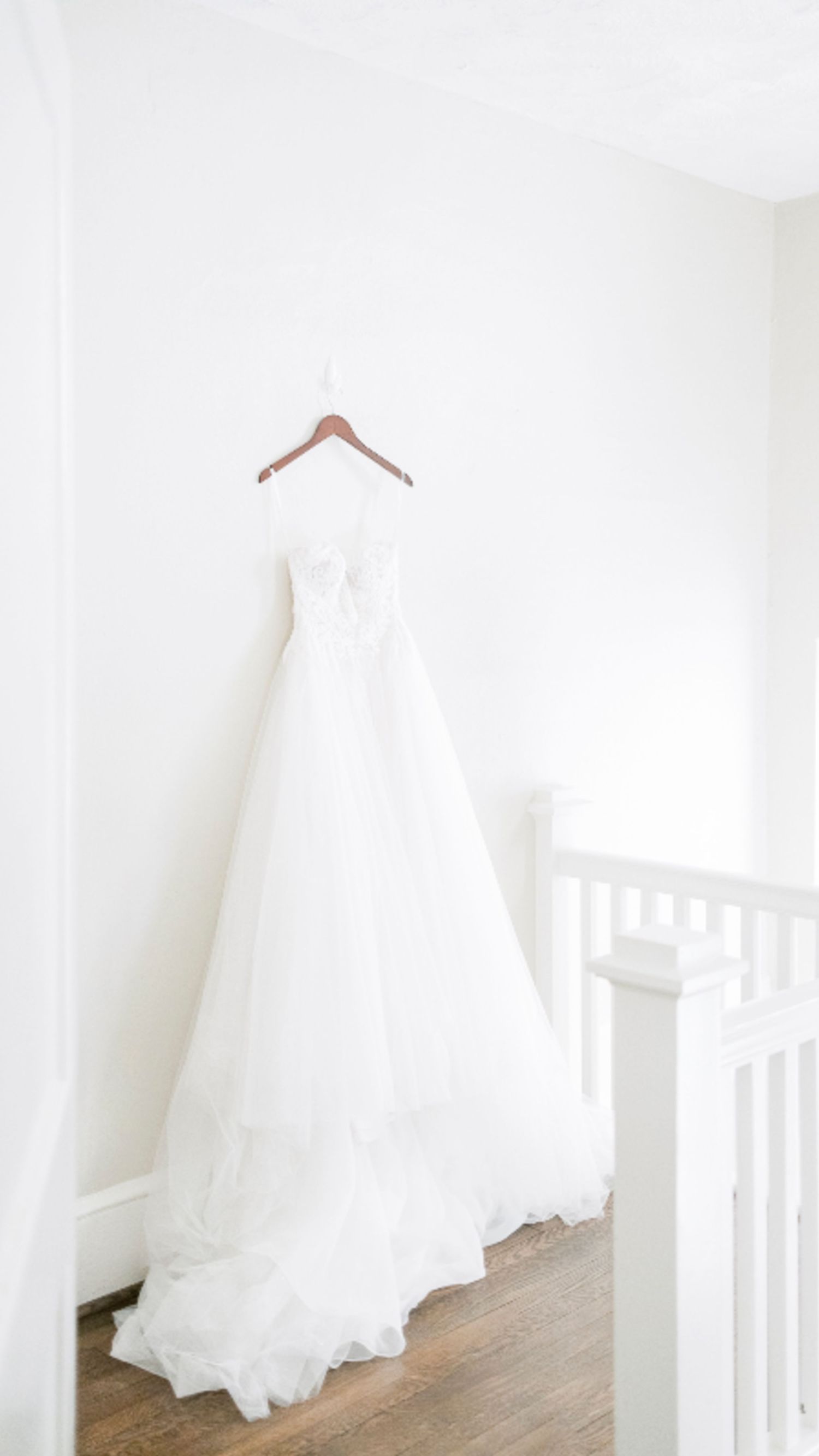 A white wedding dress hangs elegantly against a minimalist white wall, showcasing its flowing tulle skirt and delicate details.