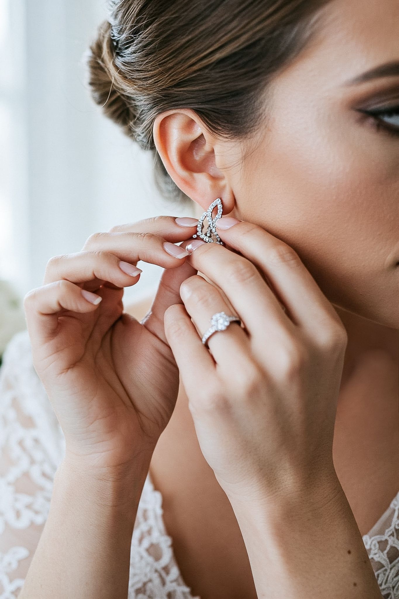 Close-up shot of hands putting on delicate earrings while wearing lace and a sparkling engagement ring.