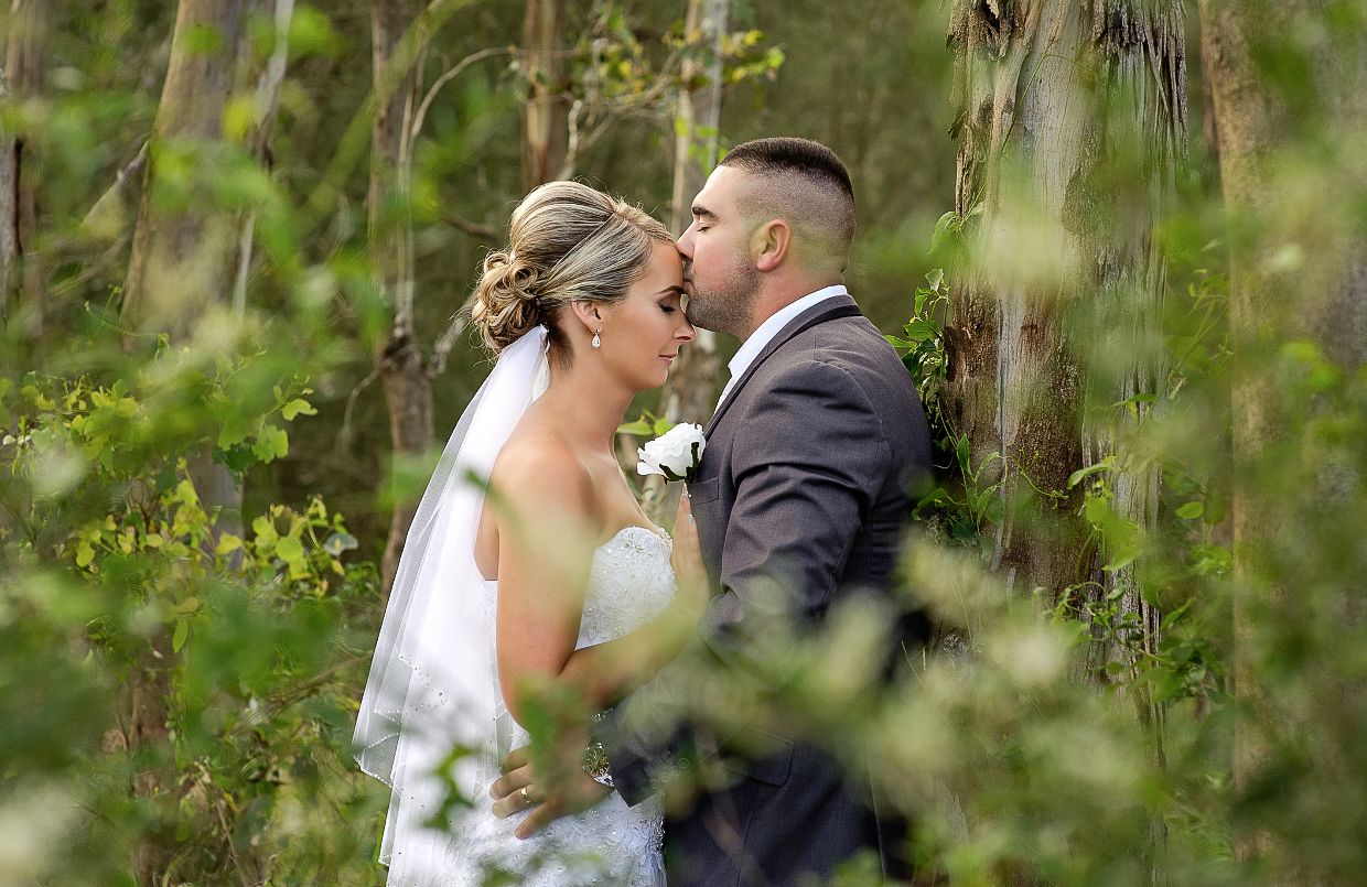 A bride and groom share an intimate moment outdoors surrounded by lush green foliage during their wedding ceremony.