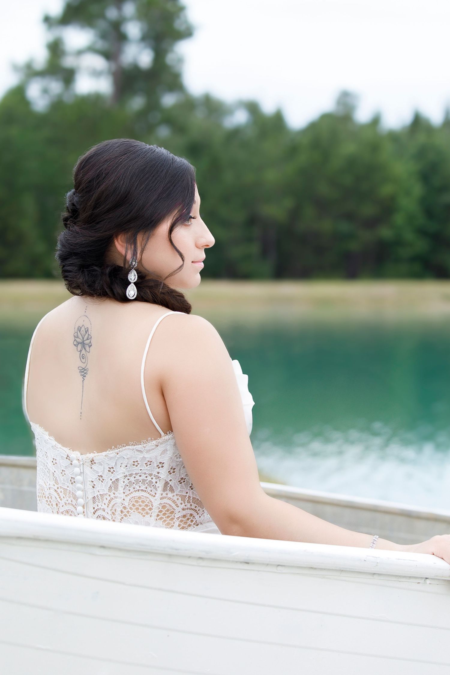 A bride in a white lace dress gazes over her shoulder while sitting on a deck by a tranquil lake.