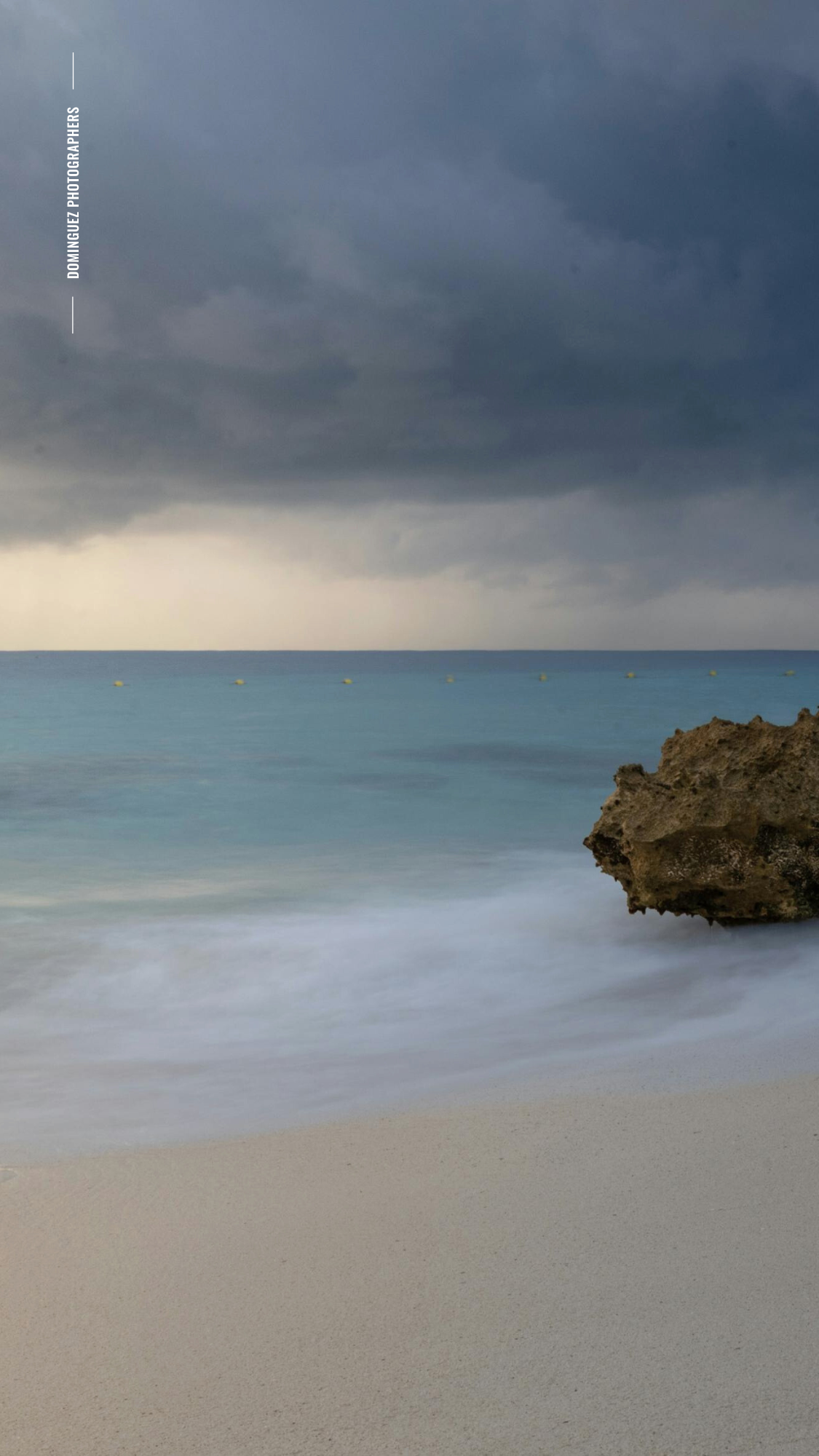 Dramatic stormy sky looms over a tranquil beach with smooth sand and rocky outcrops along the misty shoreline.
