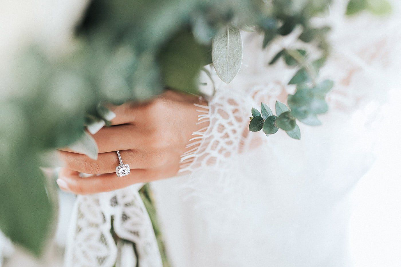 Delicate white lace wedding dress detail with floral patterns and ring on a manicured hand.