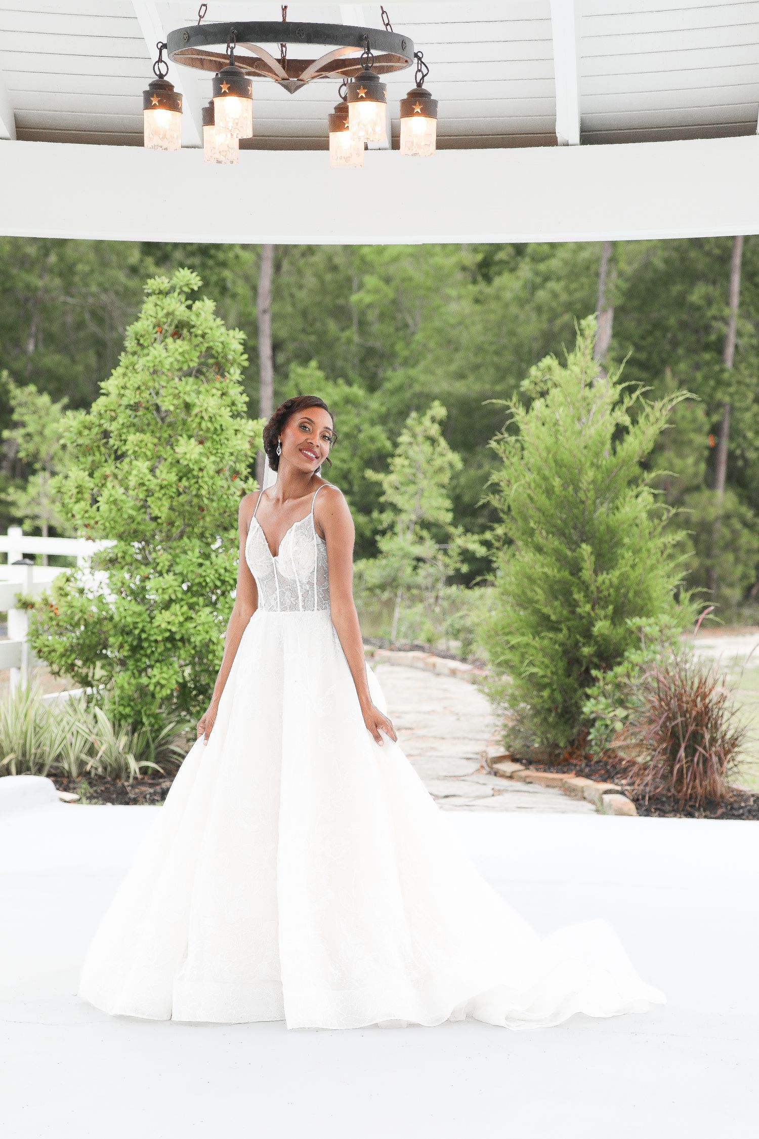 A bride in a flowing white wedding gown twirls and dances outdoors surrounded by greenery and garden landscaping.