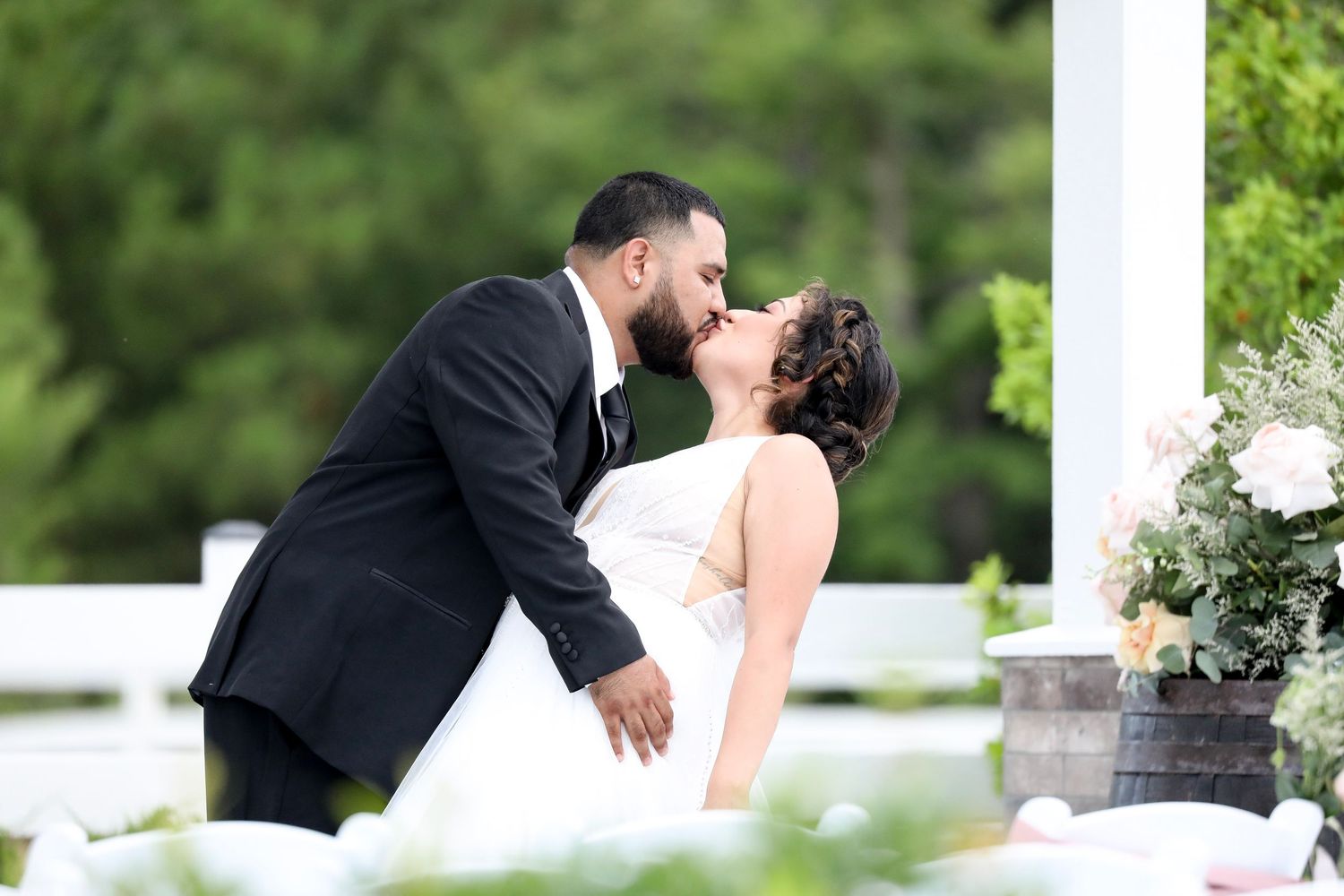 Bride and groom sharing a romantic kiss at outdoor wedding ceremony with white decor and lush green trees.