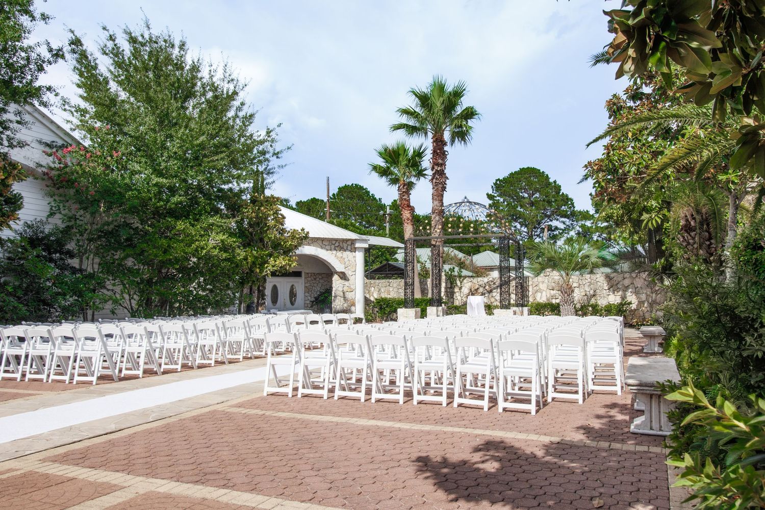 Elegant outdoor wedding ceremony setup with white chairs on brick patio surrounded by palm trees and lush greenery.