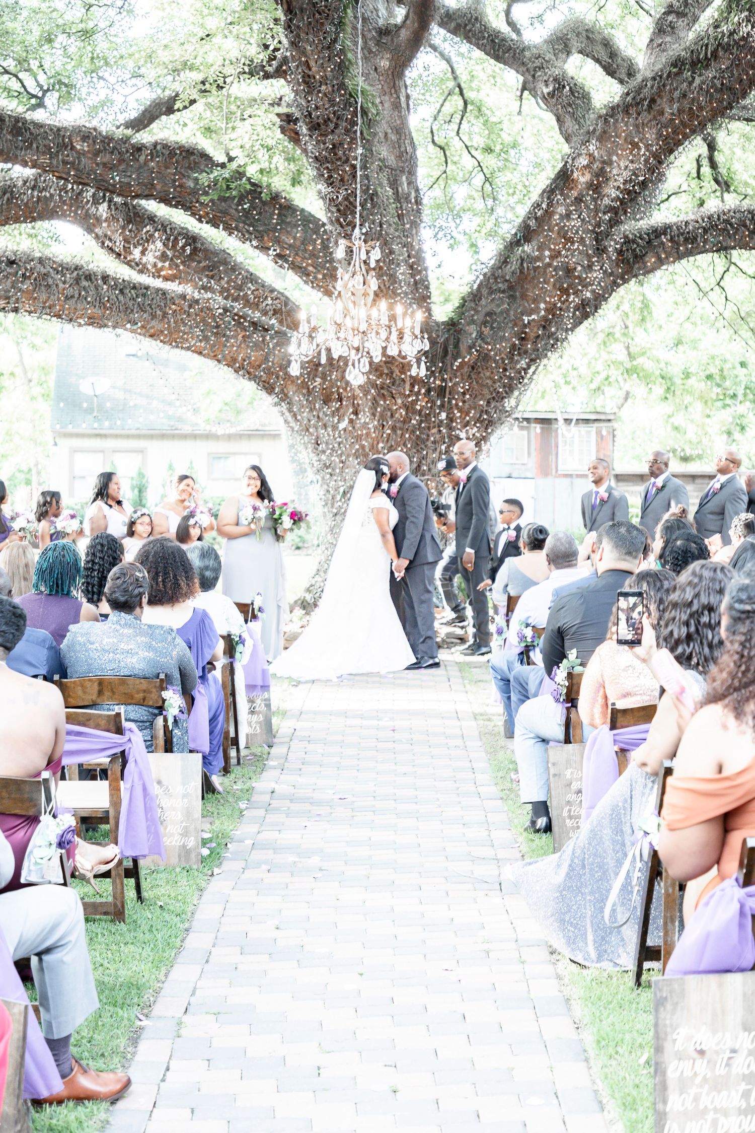 An outdoor wedding ceremony under large shade trees with guests seated in white chairs on a lawn.