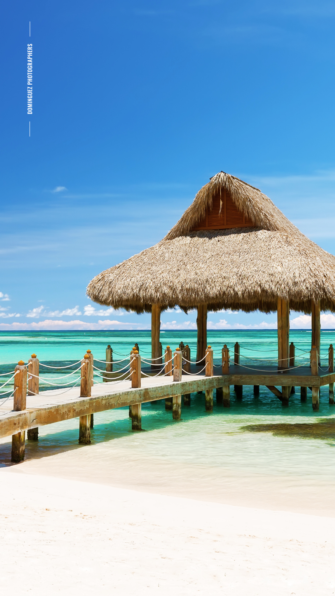 Panoramic view of thatched-roof huts on wooden stilts over crystal clear turquoise waters at a tropical beach resort.