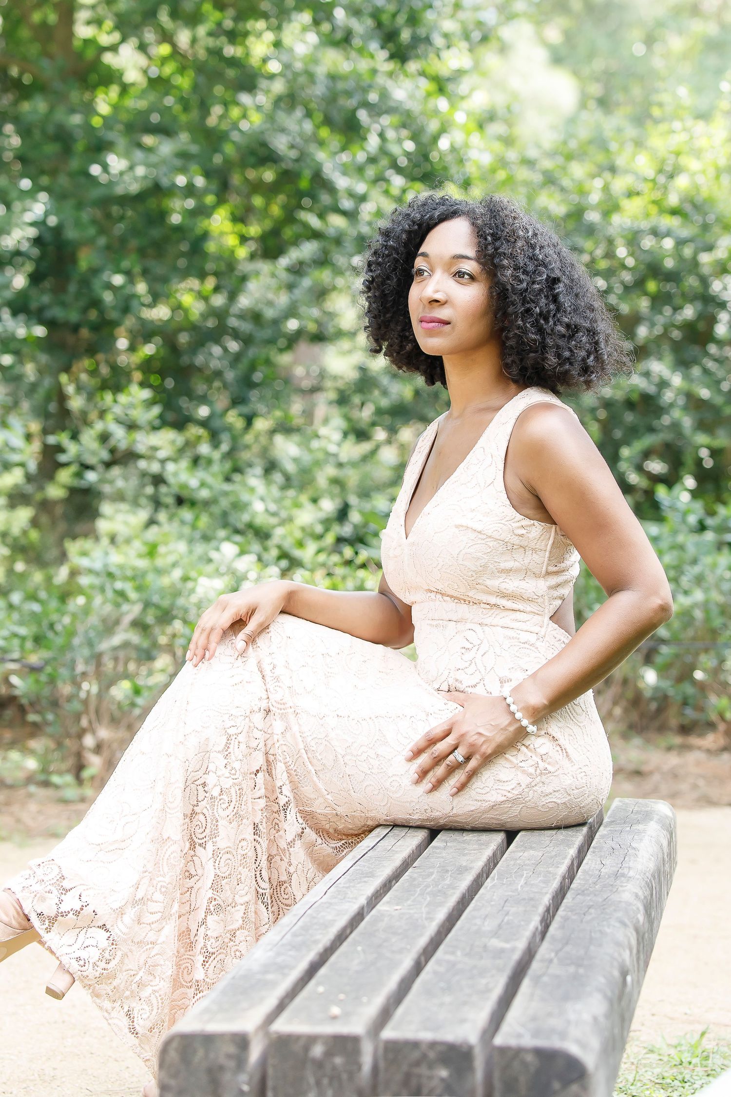 A woman in a peach lace dress poses elegantly on a wooden bench in a garden setting.