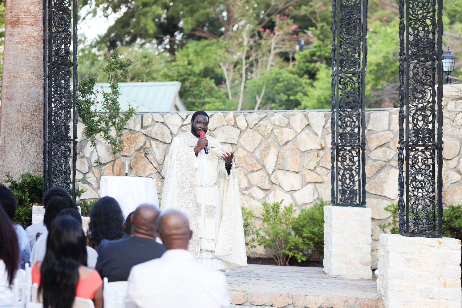 Priest in white robes speaks at an outdoor wedding ceremony beside ornate iron gates and stone wall.