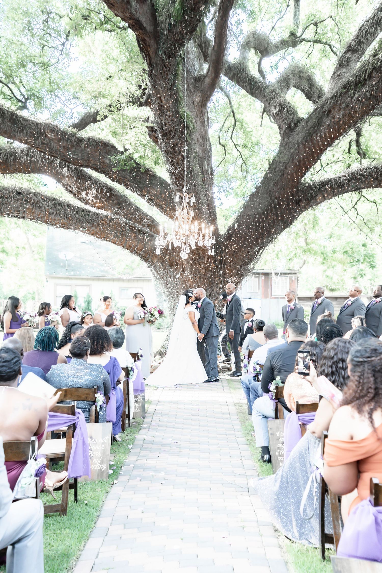 Outdoor wedding ceremony under a grand oak tree with chandelier, guests seated along aisle, bride and groom at altar.