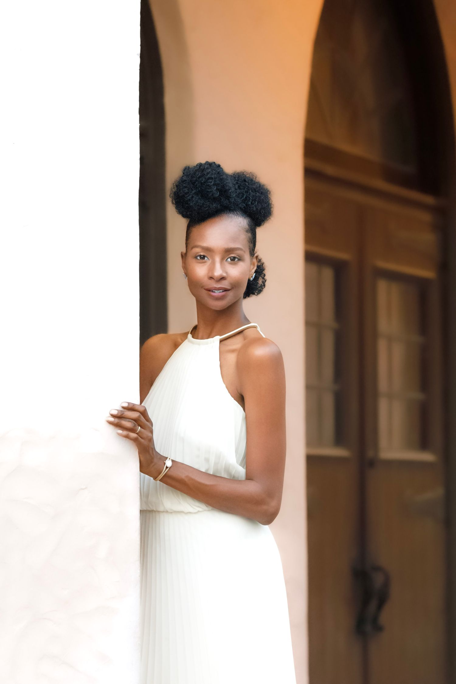 A young Black woman in a white halter dress leans against a column, smiling softly at the camera.