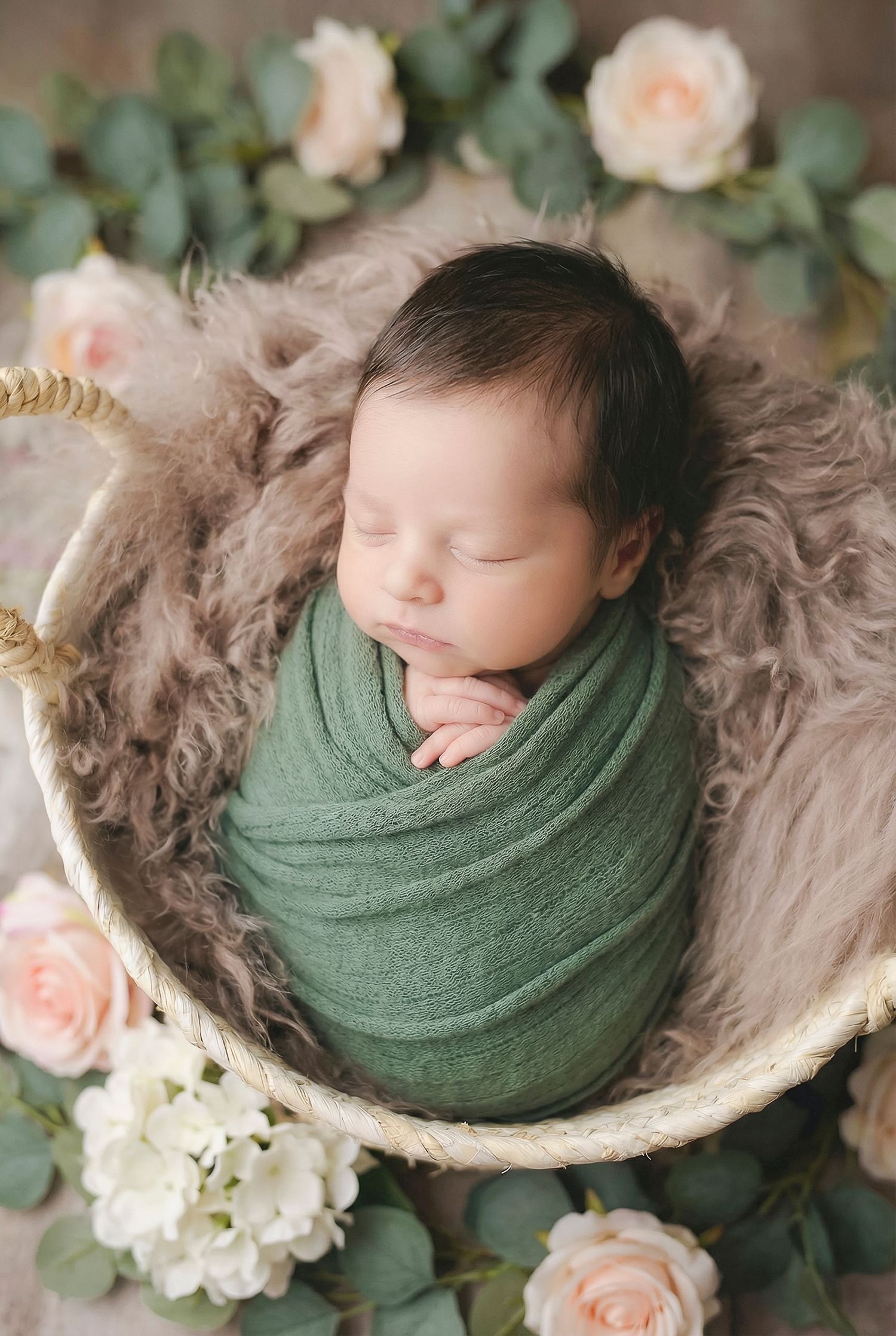 Sleeping newborn baby swaddled in green wrap nestled in a basket with soft fur, surrounded by pink and white roses.