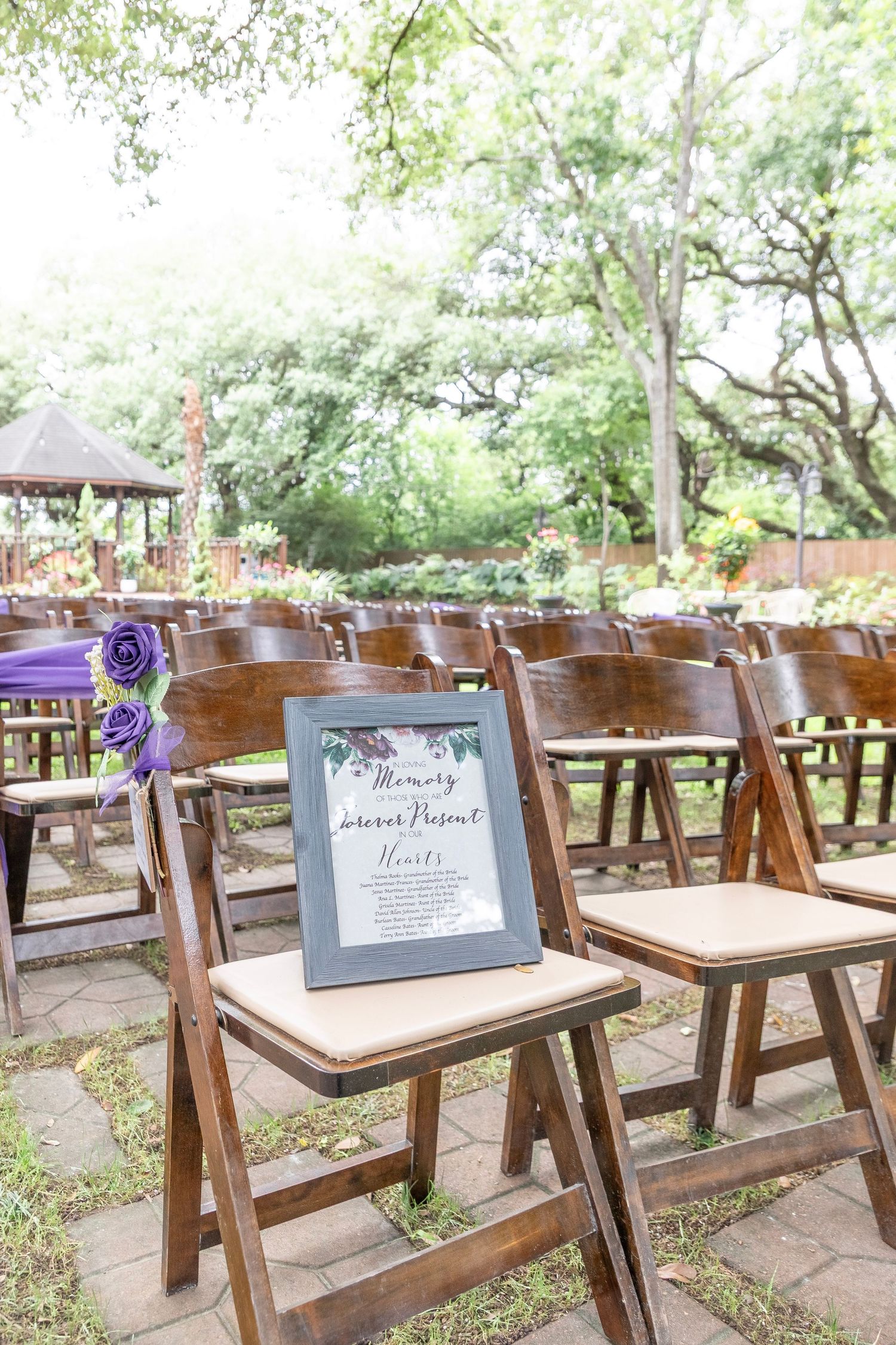Outdoor wedding ceremony setup with wooden folding chairs and a framed memorial sign in a lush garden setting.
