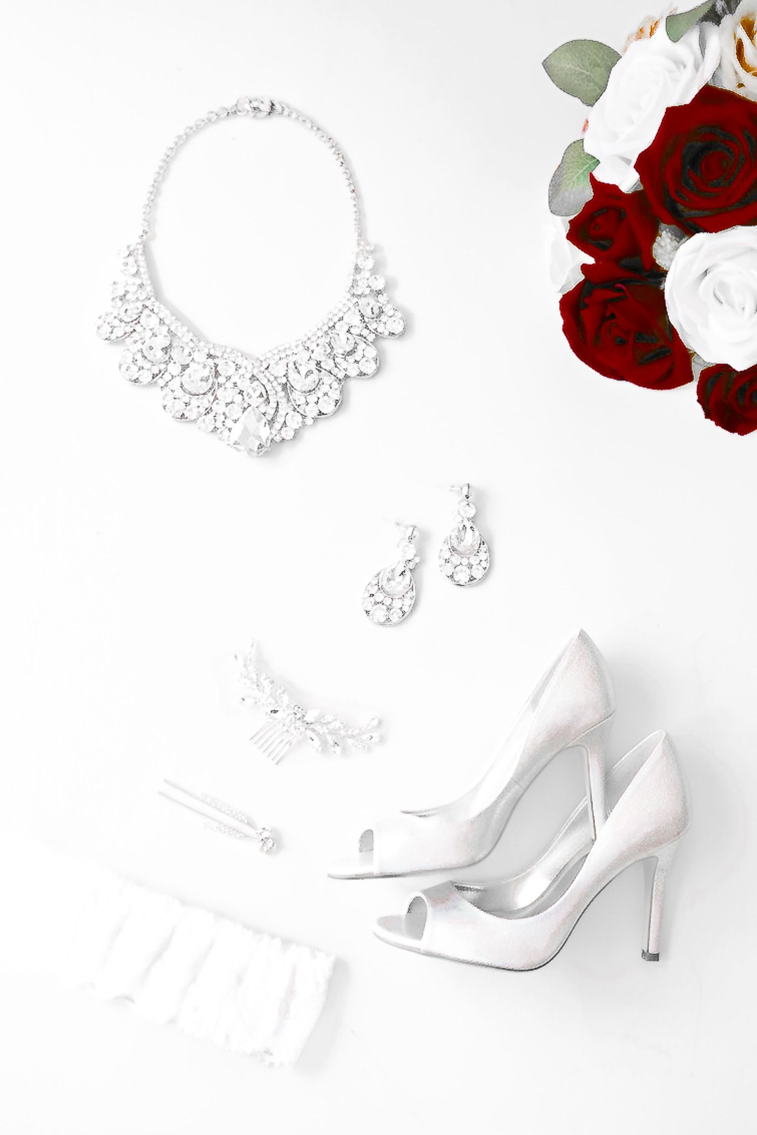 White bridal stiletto heels with crystal jewelry and red roses against a white background.